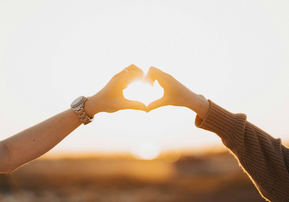 Photo qui représente deux personnes formant un coeur avec leurs mains pendant un couché de soleil.