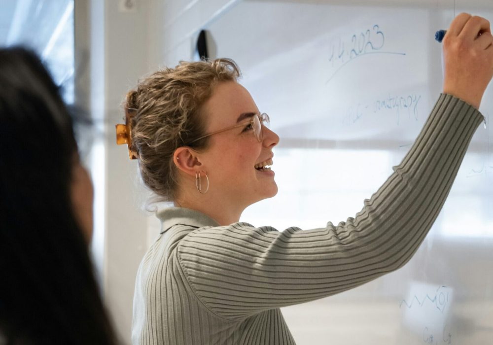 Photo qui représente une femme souriante qui écrit sur un tableau.